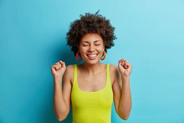 Portrait of cheerful dark skinned young woman clenches fists with triumph happy to achieve goal celebrates winning competition closes eyes dressed in casual wear isolated over blue background.