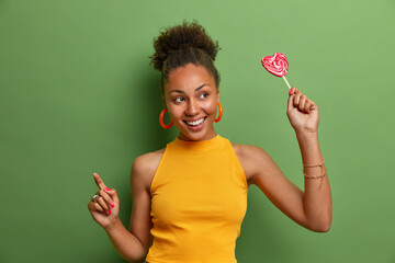 Positive beautiful curly haired woman with toothy smile holds lollipop has fun and dances carefree...