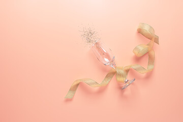 A glass of champagne with a splash of sparkles and a gold ribbon on a pastel pink background. Top view, flat lay.