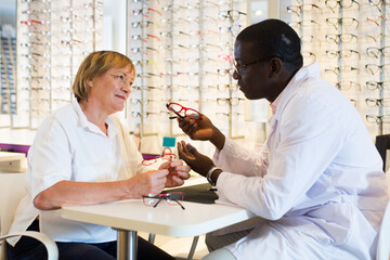 Smiling African American ophthalmologist consulting elderly woman in optical store, helping to choose eyeglasses