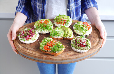 Woman holding a wooden tray with different vegan toasts with fresh microgreen sprouts.