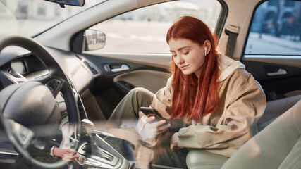 Teenage girl in earphones looking at the screen, listening to music while using her phone, sitting in the car