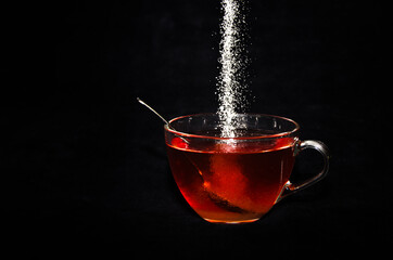 White sugar is rippling into a cup of tee in a glass, in front of black background