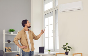 Making domestic life easy and enjoying good and convenient conditioning system: Young man standing in living-room, holding air conditioner remote control and adjusting temperature at home