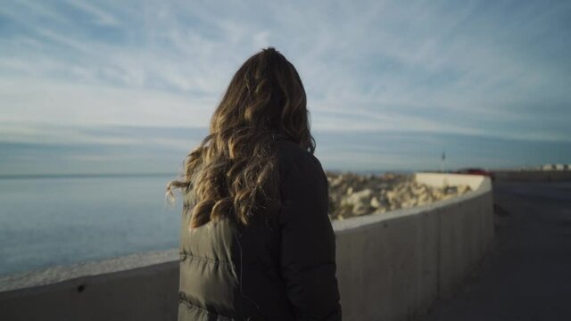 Brown haired woman walking on a black road with an amazing view of the ocean on a part cloudy day. Shot from her back, slowmo