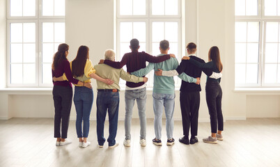 Group of diverse friends or colleagues standing together, hugging, looking out large window in new company office. Strong community of confident people, loyal business team, mutual support, friendship