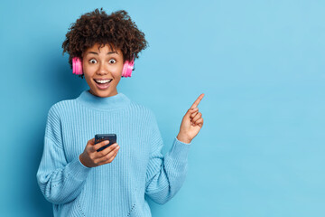 Glad ethnic woman with Afro hair indicates at upper right corner holds modern cellular listens music via stereo headphones dressed in loose knitted sweater isolated over blue background copy space