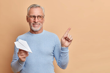 Positive bearded grey haired senior man thinks about traveling abroad holds handmade paper plane indicates at upper right corner wears casual jumper isolated over brown background shows copy space