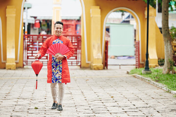 Portrait of handsome young Asian man in traditional dress for Chinese New Year standing outdoors with paper fan and red lantern - Powered by Adobe