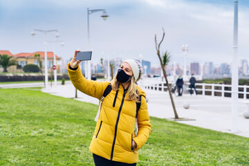 A blonde Caucasian woman in a mask uses the phone in a green part of the city. He wears a yellow coat and a woolen hat.