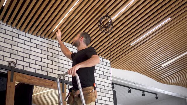 Service for the replacement of low-voltage LED lamps as well as for the repair of electrical networks. A male electrician installs stylish lamps on a wooden plank ceiling in a loft-style living room.