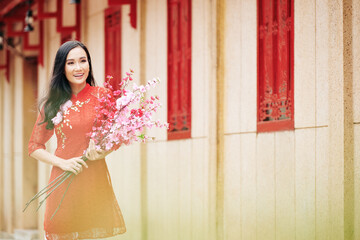 Naklejka premium Attractive smiling young Asian woman in red dress standing outdoors with blooming peach branches