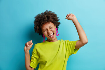 Overjoyed African American woman makes winning gesture expresses happiness raises arms and dances carefree celebrates victory dressed in t shirt isolated over blue background. Female triumphant - 406490567