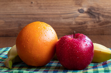 A bright red apple, orange and banana lie on a linen towel. There are drops of water on the fruit. In the background is a wooden wall. Fruits are taken in close-up.