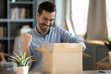 Happy 30s young man in eyewear looking inside of carton box, celebrating getting wished item or...