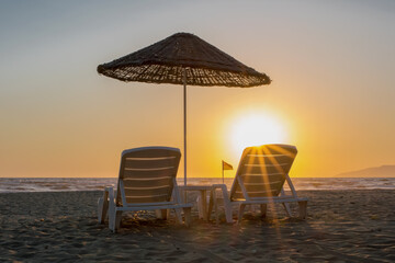 Two free recliners on beach sand under big wooden umbrella near sea water at sunset.