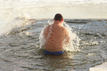 Winter ice swimming sport, a European man in a swimming trunks swimm in the ice hole water with splashes on a Sunny frosty winter day, healthy lifestyle
