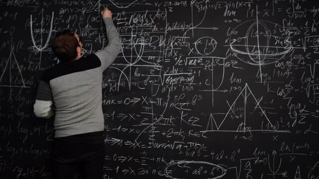 Professor writes equation on blackboard full of mathematics formulas and points finger smiling. Smart teacher explains material. A young caucasian man solves problem. Concept of science and education.