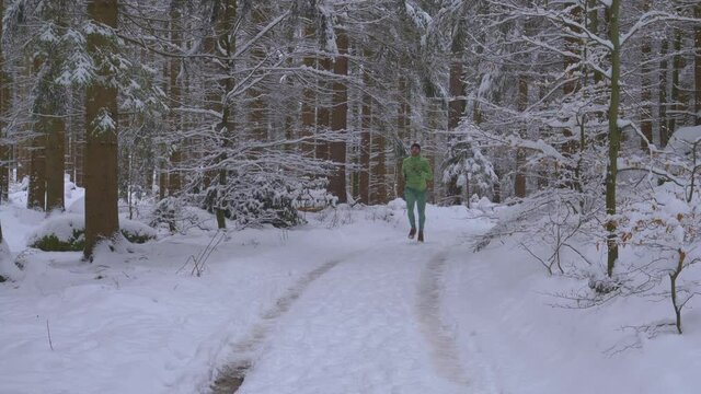 Athlethic Adult Man Running From Distance In Green Outfit In Snow Covered Forest On Wild Path During Cold, Winter Day - Toward Camera, Slow Motion