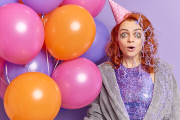 Fototapeta na wymiar Horizontal shot of surprised foxy young woman stares at camera keeps mouth opened smeared with cake cream wears party hat holds bunch of multicolored balloons isolated over purple background