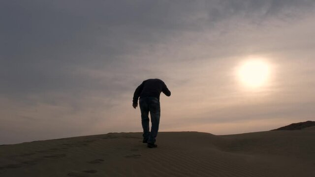 A man lost in the desert stumbles across the sand into the sunset.