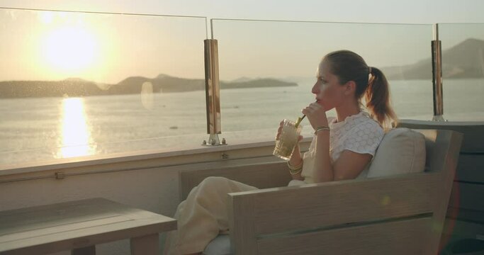 good looking woman sitting on a chair on a rooftop drinking a cocktail with a straw, with a beautiful sunset above the sea in the background