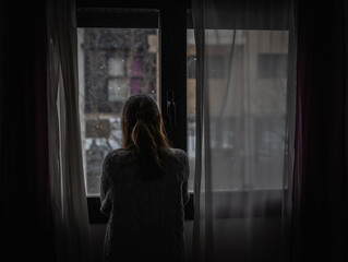 Young woman looks out a window between the curtains while it is snowing