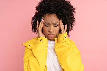 Obraz na płótnie Canvas Unhappy Afro woman with curly hair in wear yellow raincoat suffering from severe throbbing headache, touching her temples and head, closed eyes, isolated on studio pink background