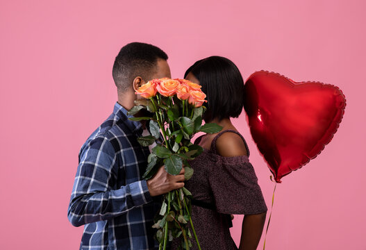 Loving African American couple kissing behind bunch of roses, holding heart shaped balloon on pink studio background