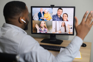 Virtual meeting with a family, friends. View over an African guy shoulder on the laptop screen with a multigenerational people on the screen, senior couple and diverse young people talking via vide