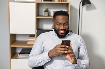 Biracial guy in smart casual shirt is using smartphone in the office. An African-American employee chatting, texting messages, scrolling news feed on the cellphone on a workplace