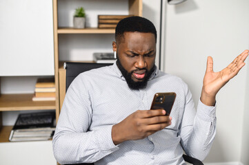 Confused puzzled African-American man in smart casual shirt holding smartphone and expressing misunderstanding, spreads his hands in incomprehension - obrazy, fototapety, plakaty