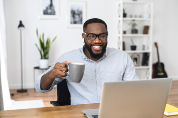 Fototapeta na wymiar Smiling African-American businessman in glasses sitting at the desk, working from home, holding a cup of coffee and laughing, looking at the laptop screen, and participating in the webinar