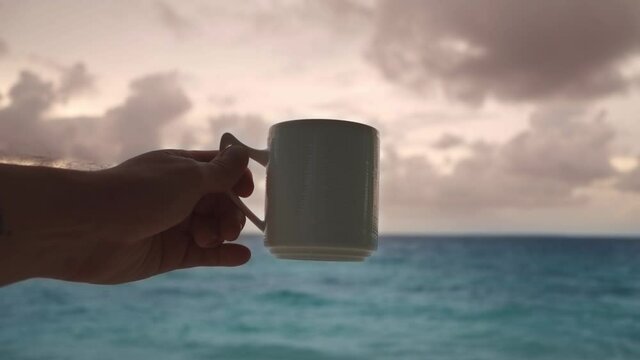 A man is holding a large mug of hot chocolate or coffee, or maybe tea - a close-up against the backdrop of a beautiful sunrise at the sea.
