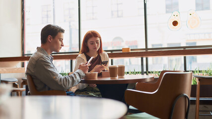 A couple of teenagers totally absorbed in using their phones, ignoring each other while sitting in a cafe together on a daytime