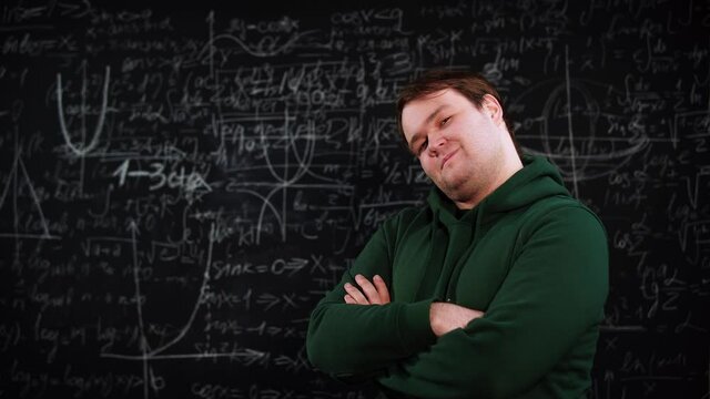 Portrait of a satisfied student standing in front of a chalkboard where there are algebra formulas. He smiles and crosses his hands on chest. A young man is glad of having solved mathematics equations