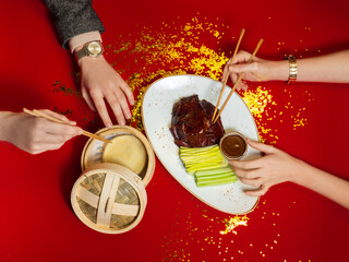 Chinese national dish Peking duck. Male and female hands reaching for food. In the hands of Chinese sticks. Dinner for two. Asian food stands on a red background, with golden lines scattered nearby.