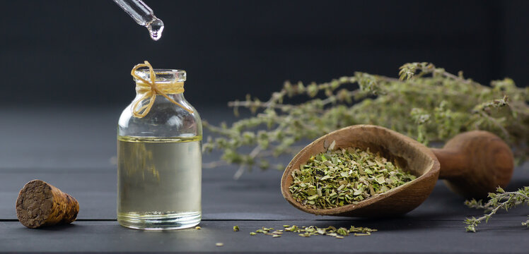 Glass bottle of thyme essential oil and bunch of dry thyme on black wooden rustic background, oil dripping