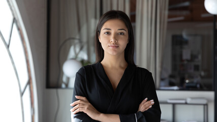 Head shot portrait confident Indian businesswoman hr manager standing in modern office with arms crossed, serious entrepreneur team leader mentor posing for corporate photo, looking at camera