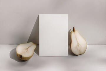Trendy summer stationery still life scene. Closeup of cut pear fruit on beige table background in sunlight. Blank paper card, invitation mockup scene lean on champagne wall, long shadows.