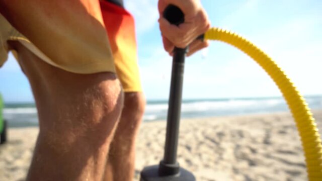 Guy on a beach in colorful boardshorts using a hand pump to inflate a kite while someone is kitesurfing in the background