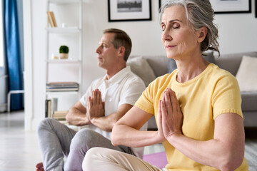 Calm healthy serene old middle aged senior 50s couple meditating with eyes closed namaste hands at home together. Senior retired man and woman doing yoga exercise, breathing, feeling no stress, zen.