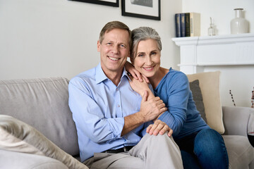Happy older 50s family couple hugging, looking at camera, sitting on couch in modern apartment. Smiling affectionate senior middle aged european husband and wife embracing posing for portrait at home.