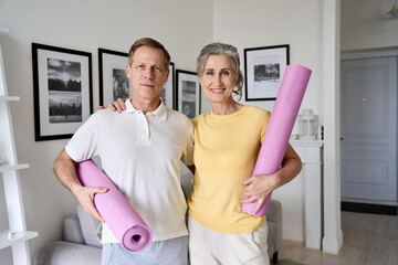 Happy fit middle aged old couple hugging, looking at camera, holding yoga mats, standing at home. Seniors fitness exercises, health and body care training, mature people active lifestyle. Portrait.