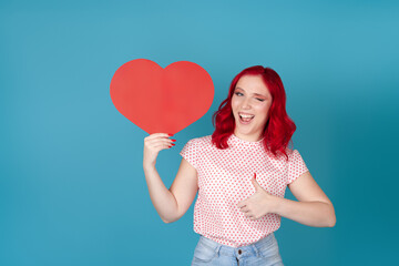 portrait of a mischievous, playful young woman with red hair holding a red paper heart, winking and giving a thumbs up, isolated on a blue background.