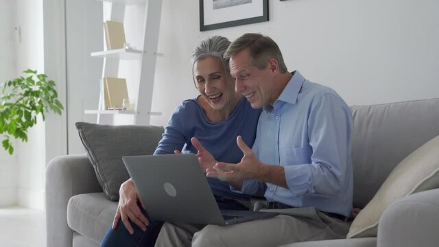 Happy senior couple talking looking at laptop making online video call. Middle aged old grandparents enjoying virtual distance meeting online chat looking at computer sitting on couch at home.