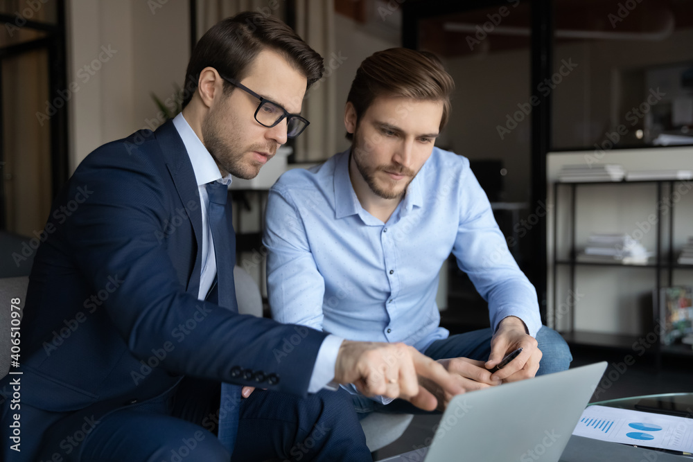 Poster close up serious mentor wearing glasses teaching new employee, intern, pointing at laptop screen, si