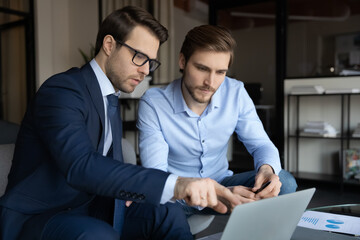 Close up serious mentor wearing glasses teaching new employee, intern, pointing at laptop screen,...