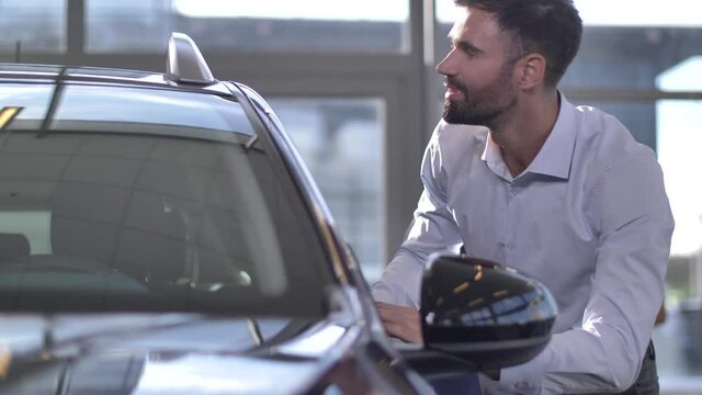 Close-up of joyful male buyer looking at new luxury car chosen for purchase with delight while visiting auto dealership, at same time his wife conversating with salesman blurry on background