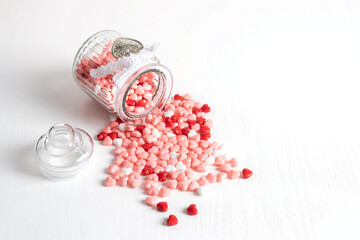 Valentine day background. Multi-colored little heart-shaped candies pour out of a glass jar. White lace braid with a metal heart.